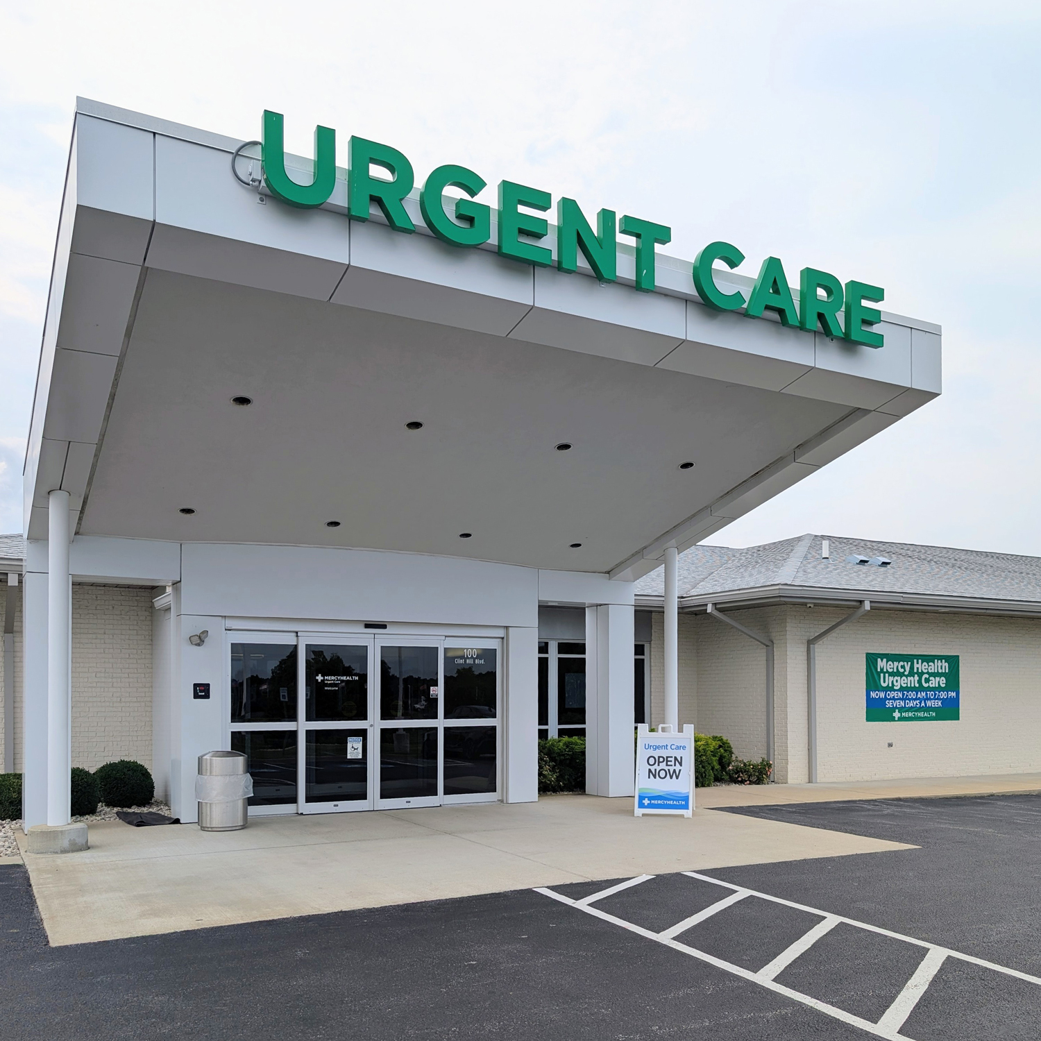 Exterior of Mercy Health Urgent Care in Paducah, KY, showing the entrance under a large canopy with a green 'Urgent Care' sign, an 'Open Now' sidewalk sign, and the parking lot in the foreground